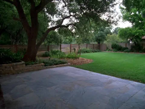 Shaded backyard patio under a live oak with a stacked-limestone planter and native beds.